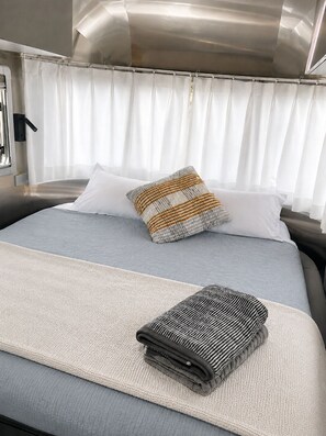 Un lit avec un oreiller à rayures grises et blanches, une couverture pliée et un rideau blanc.