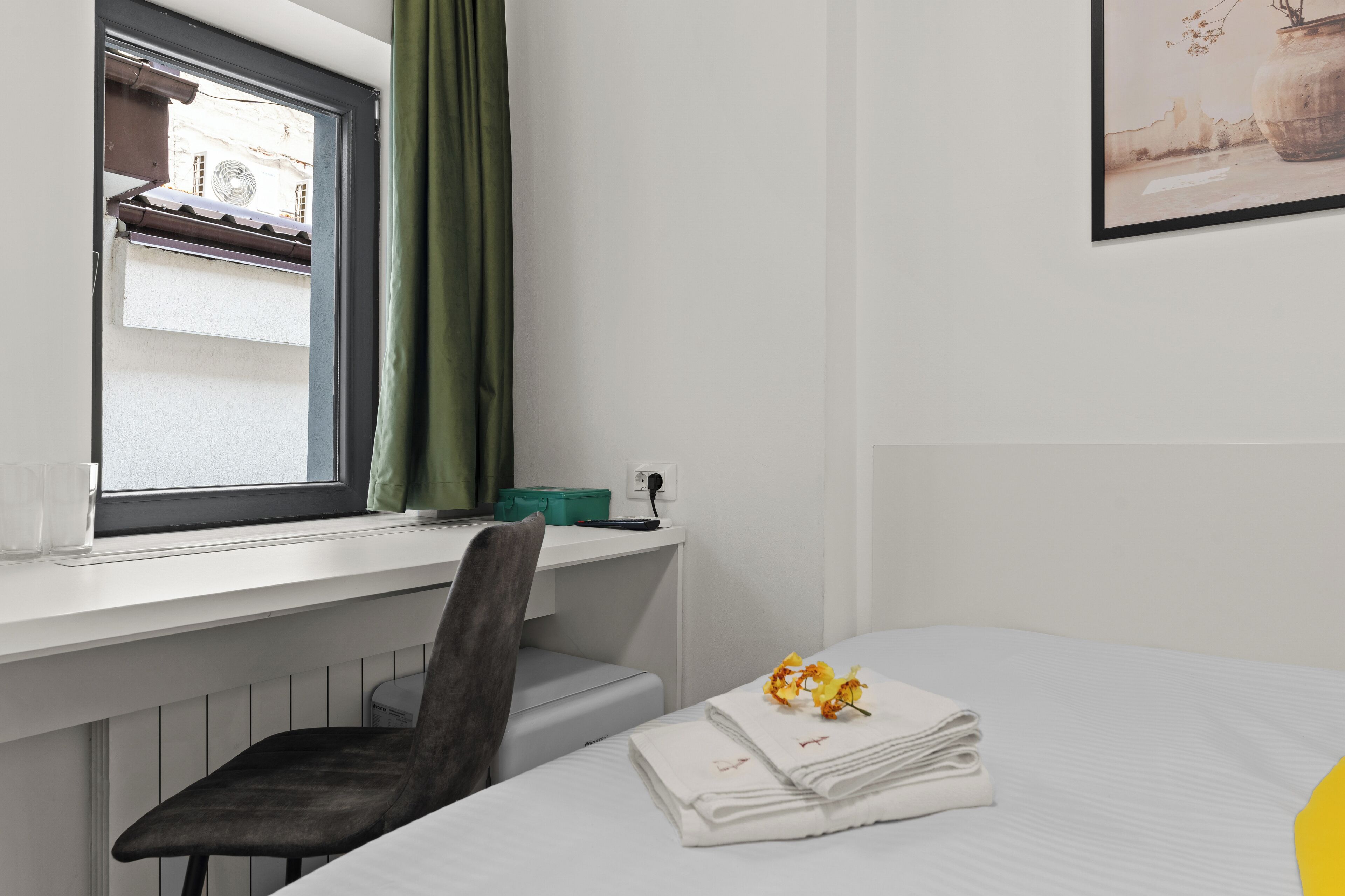 Habitación de hotel con cama, escritorio con silla, ventana con vista a edificios y un cuadro en la pared.
