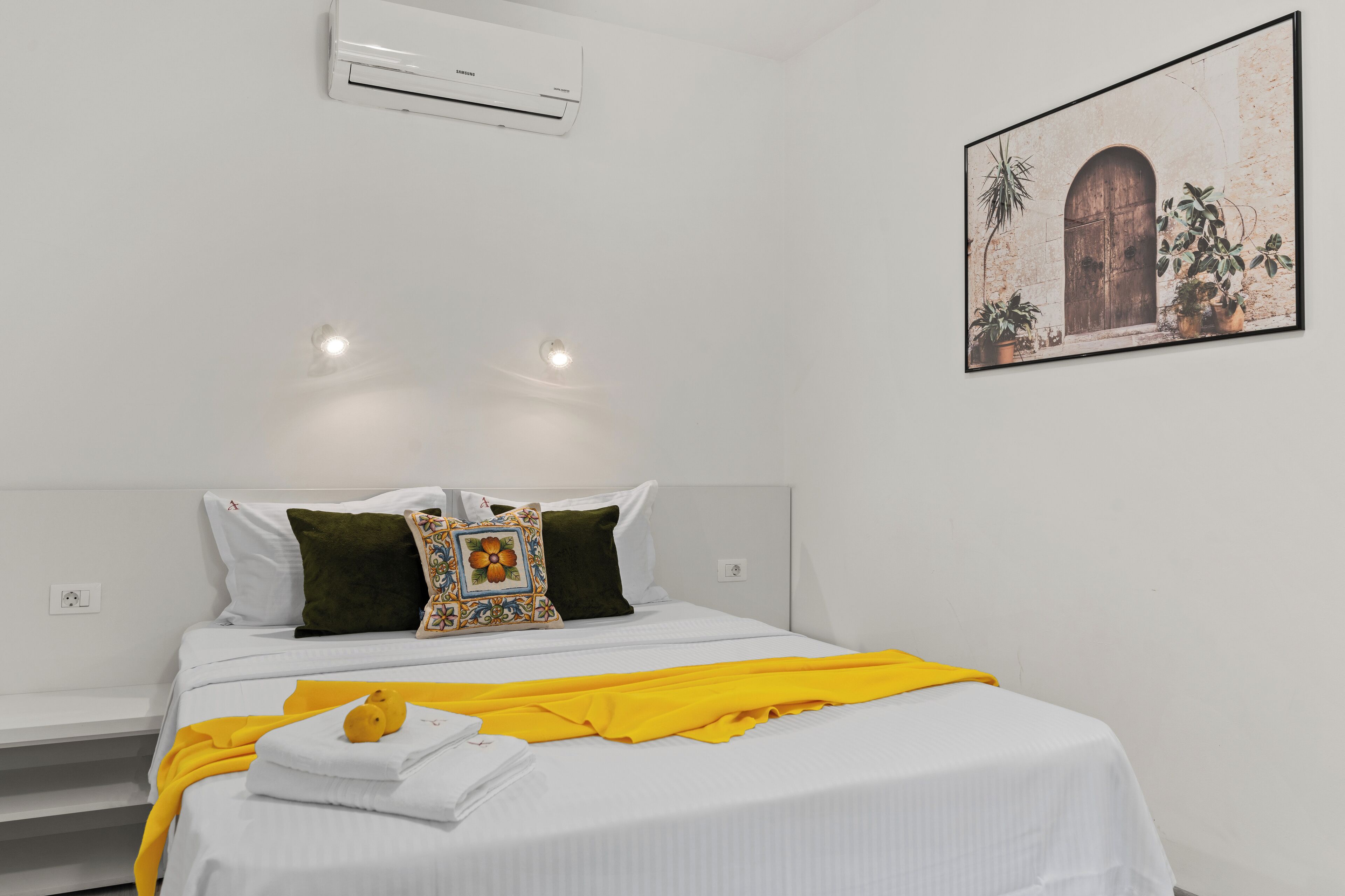 Een slaapkamer met een bed, witte beddengoed, een gele plaid en een luchtverkoeler aan de muur.