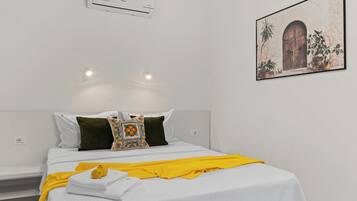 Een slaapkamer met een bed, witte beddengoed, een gele plaid en een luchtverkoeler aan de muur.
