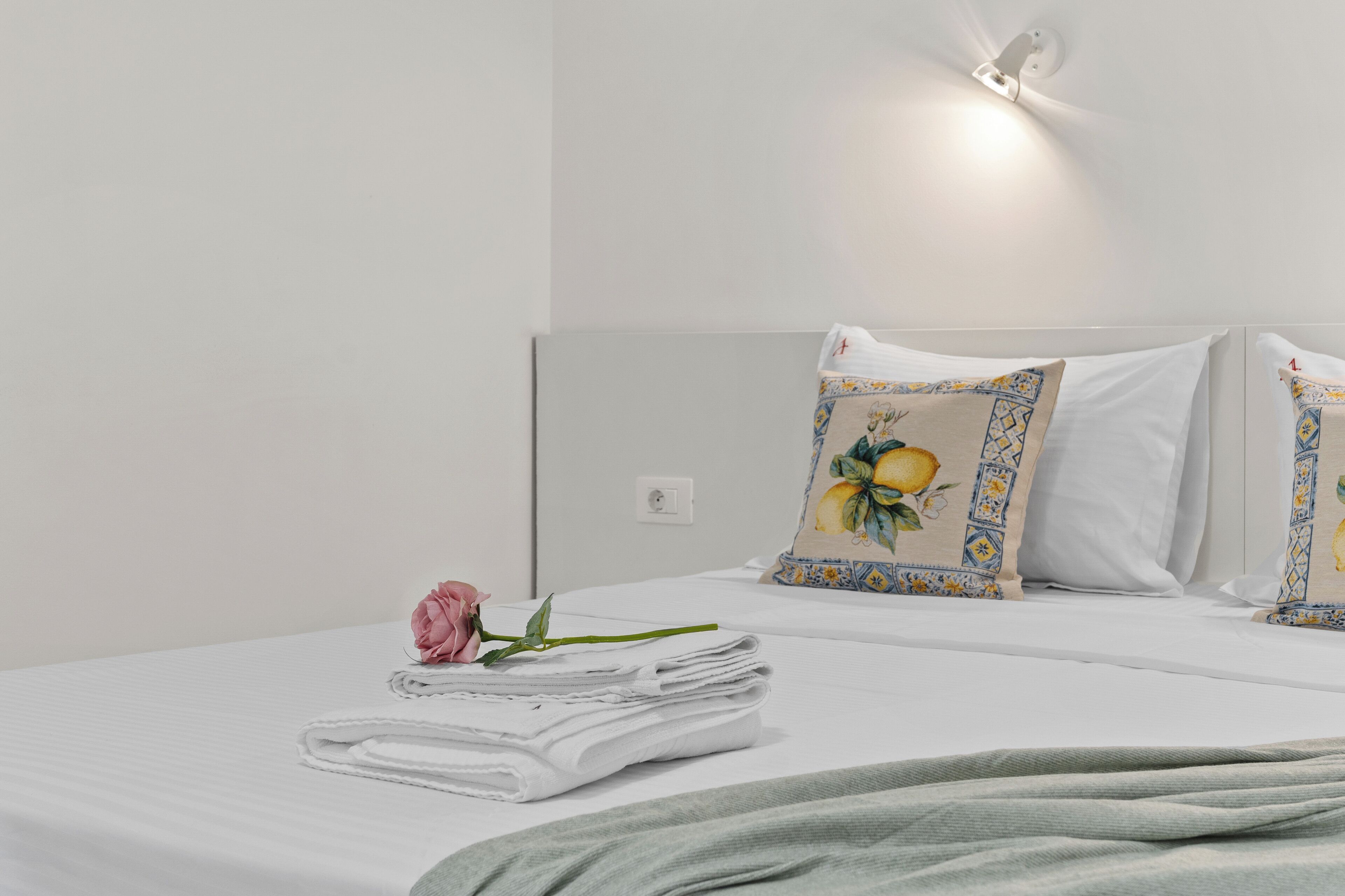 A neatly made bed with a folded towel, a pink rose, and decorative pillows with a fruit pattern.