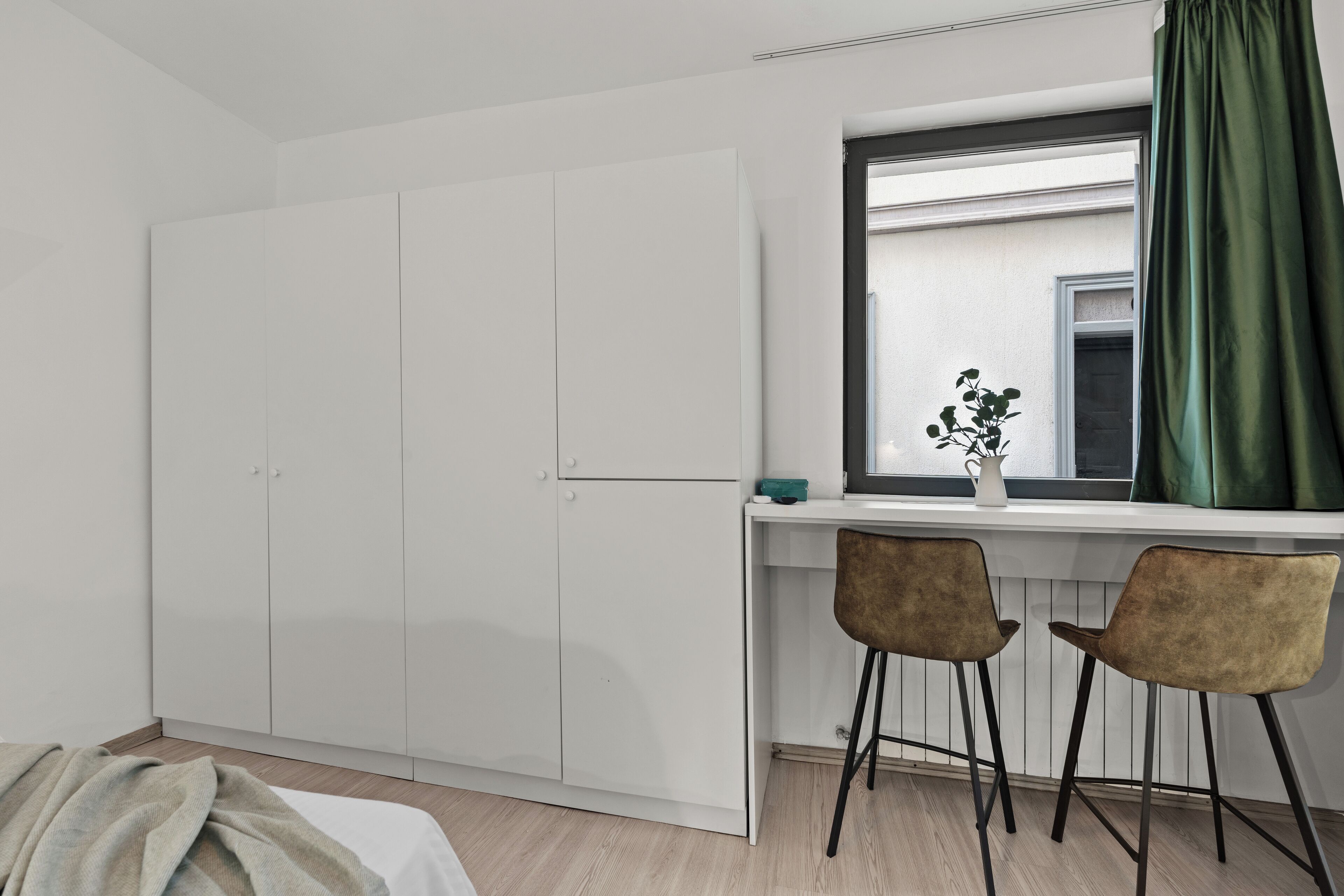 Um quarto com cama, guarda-roupa branco, escrivaninha com duas cadeiras, janela com cortina verde e planta em vaso no parapeito.