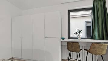 Um quarto com cama, guarda-roupa branco, escrivaninha com duas cadeiras, janela com cortina verde e planta em vaso no parapeito.