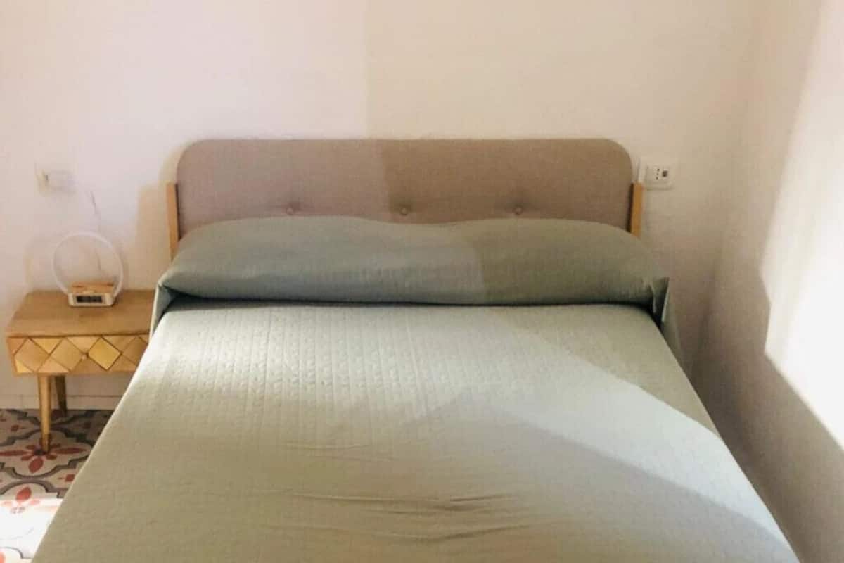 A single bed with a headboard, a bedside table with a lamp, and a power outlet on the wall.
