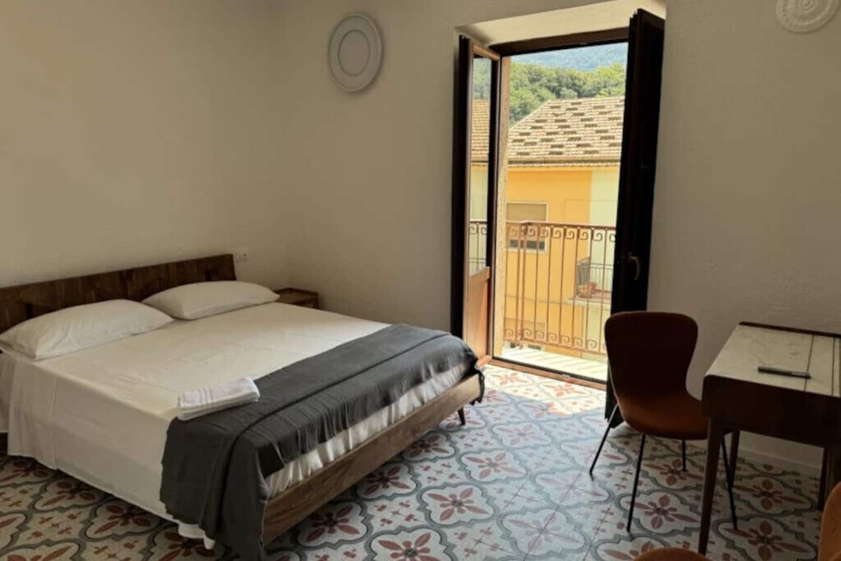 A bedroom with a bed, a table, a chair, and a balcony with a view of the outside.