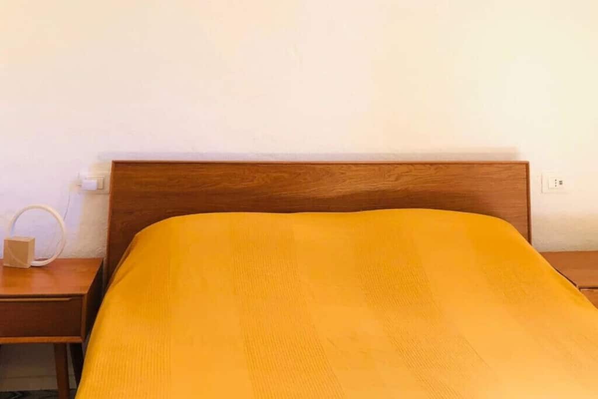 A bed with a wooden headboard, a bedside table with a lamp, and a wall socket.