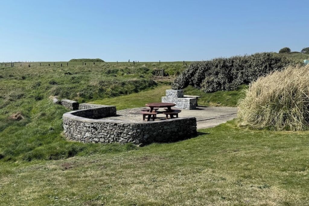 A picnic area with stone benches and a picnic table, surrounded by grassy terrain and a stone wall.