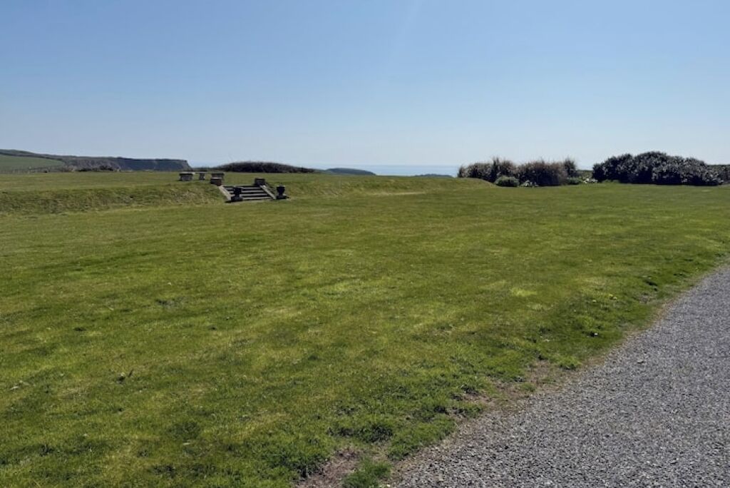 A grassy field with a gravel path, benches, and a distant view of the sea.