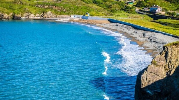 Blick auf die Küste mit klarem, blauen Wasser, einer felsigen Uferlinie und einer Grünfläche mit Gebäuden.