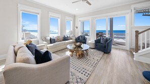 A spacious living room with a large window overlooking the ocean, a ceiling fan, and a wooden staircase. - Joy By The Sea 5 Bedroom Home (Panama City Beach)