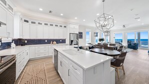 A modern kitchen with white cabinetry, a large island, and a dining area with a view of the ocean. - Joy By The Sea 5 Bedroom Home (Panama City Beach)