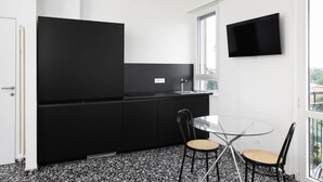 Ett modernt kök med svarta skåp, ett glasbord och stolar, en platt-TV som är monterad på väggen och ett fönster som släpper in naturligt ljus.