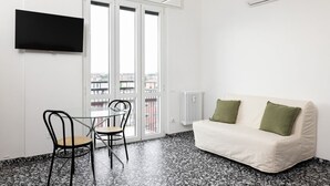Ett rum med en vit soffa, två gröna kuddar, en svart tv som är monterad på väggen, ett glasbord med två stolar och ett stort fönster med utsikt över staden.