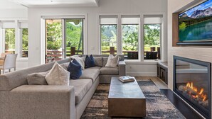 A modern living room with a sectional sofa, a fireplace, large windows with a view of greenery, and a flat-screen TV mounted on the wall. - Chaparral 204 3 Bedroom Condo (Park City)