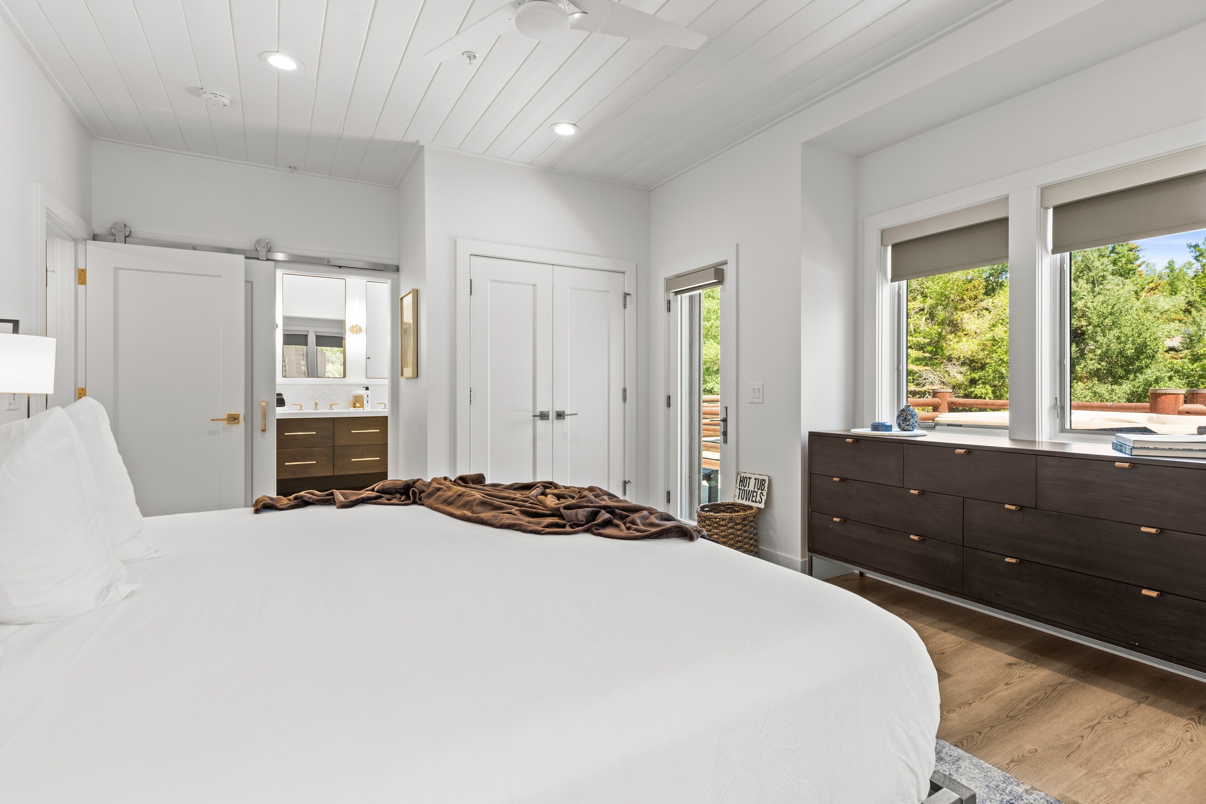 A bedroom with a large bed, a dresser, a mirror, and a window with a view of greenery.