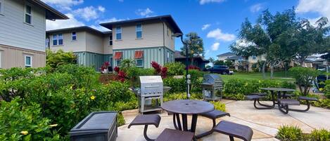 An outdoor patio with picnic tables and a barbecue grill, surrounded by lush greenery and residential buildings.