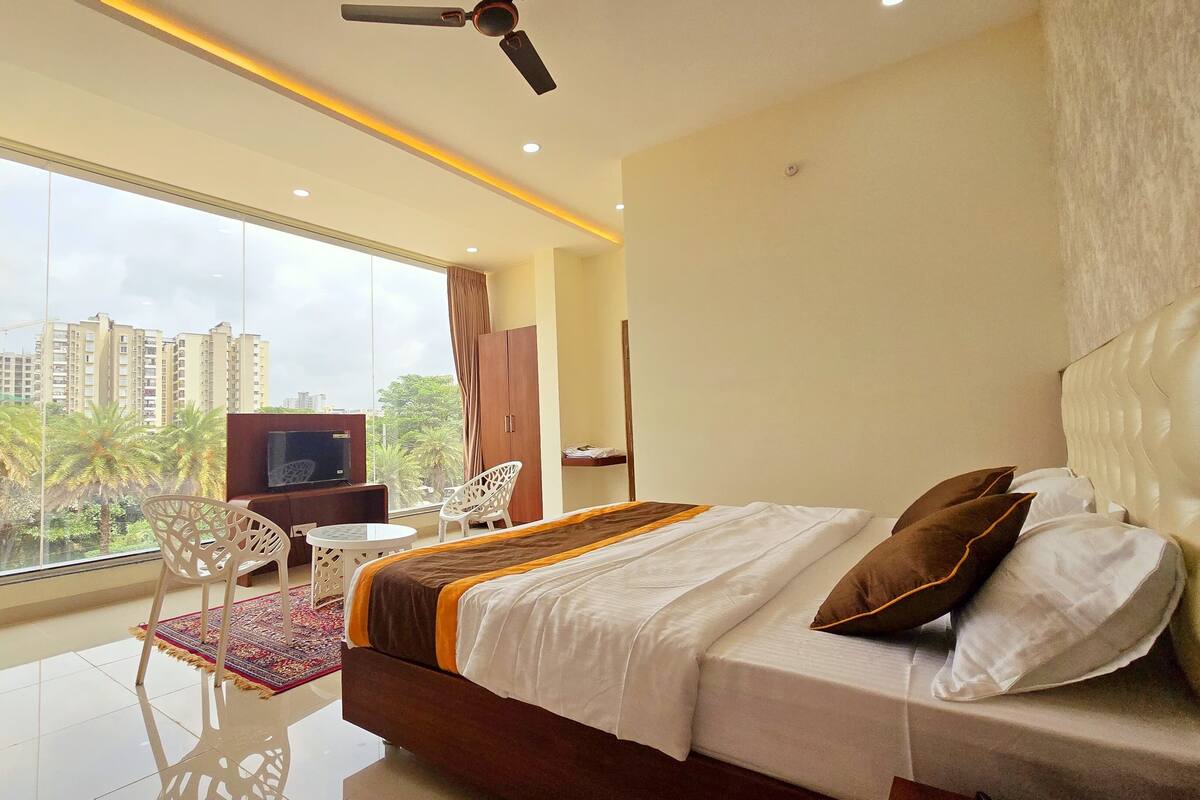 A hotel room with a large bed, a TV, a desk, and a view of palm trees and buildings.