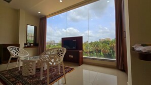 Habitación de hotel con un ventanal grande, un televisor, dos sillas blancas, una mesita y una alfombra estampada.