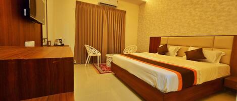 A hotel room with a wooden desk, a bed with a headboard, a TV mounted on the wall, and an air conditioning unit.