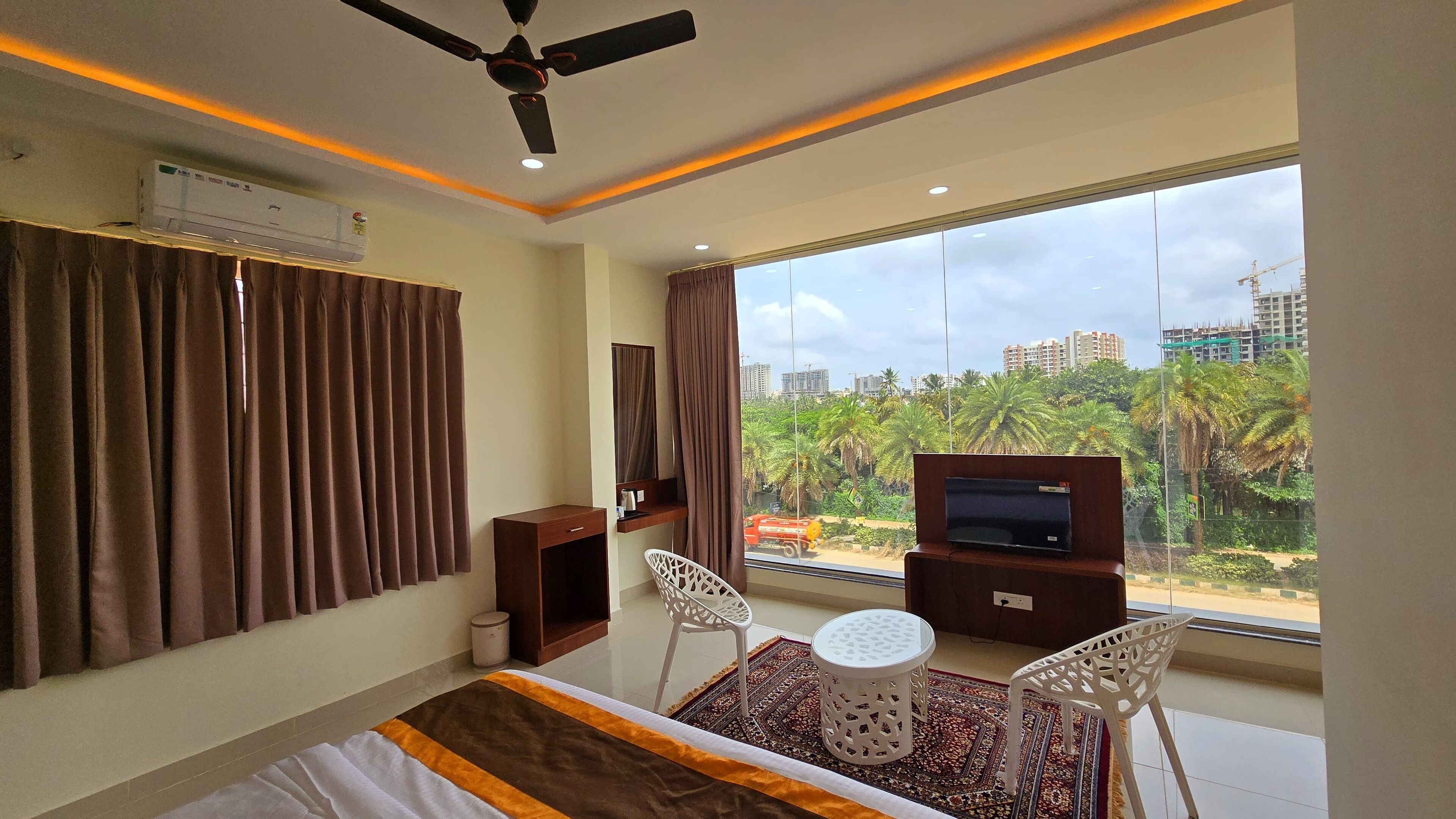 A hotel room with a large window, a ceiling fan, an air conditioning unit, a TV, a small table, two chairs, and a bed with a patterned bedspread.