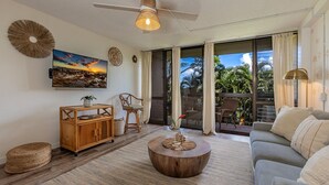 A living room with a wooden coffee table, a sofa, a TV mounted on the wall, and a ceiling fan. - Maui Vista 2223 1 Bedroom Condo (Kihei)