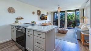 A modern kitchen with a large island, a dining area with a sofa, and a view of greenery through the sliding glass doors. - Maui Vista 2223 1 Bedroom Condo (Kihei)