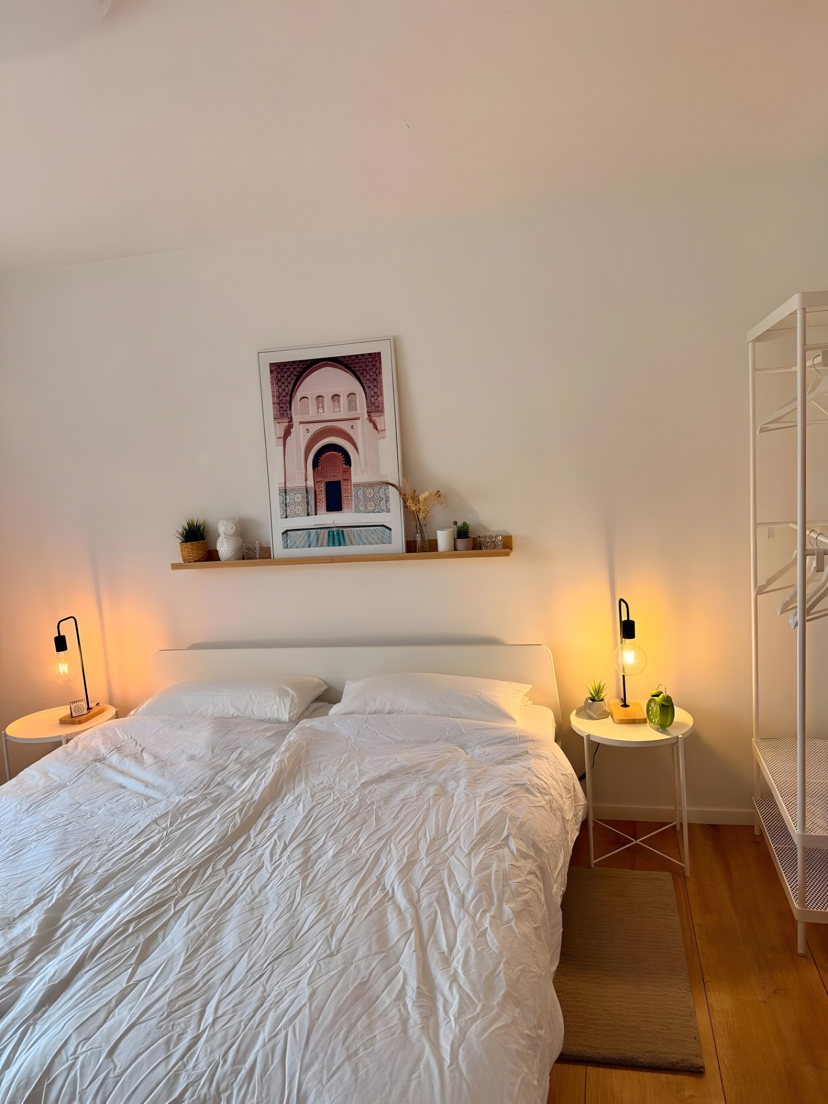 A bedroom with a large bed, two bedside tables with lamps, a wall-mounted shelf with decorative items, and a bunk bed structure.