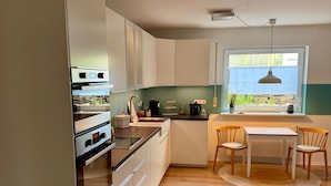 Une cuisine moderne avec des meubles blancs, un four encastré, une table à manger avec des chaises et une fenêtre donnant sur un beau paysage.