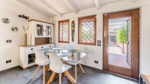 Un coin repas avec une table et des chaises en verre, un meuble blanc aux portes en verre et une porte en bois donnant sur l’extérieur.