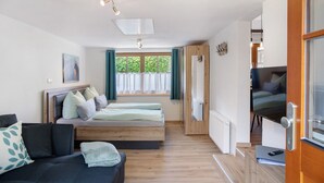A compact room with a bed, a sofa, a window with curtains, a door leading to another room, and a small table with a remote control. - Holiday Apartment 'Wankblick' with Mountain View, Private Terrace and Wi-Fi (Farchant)