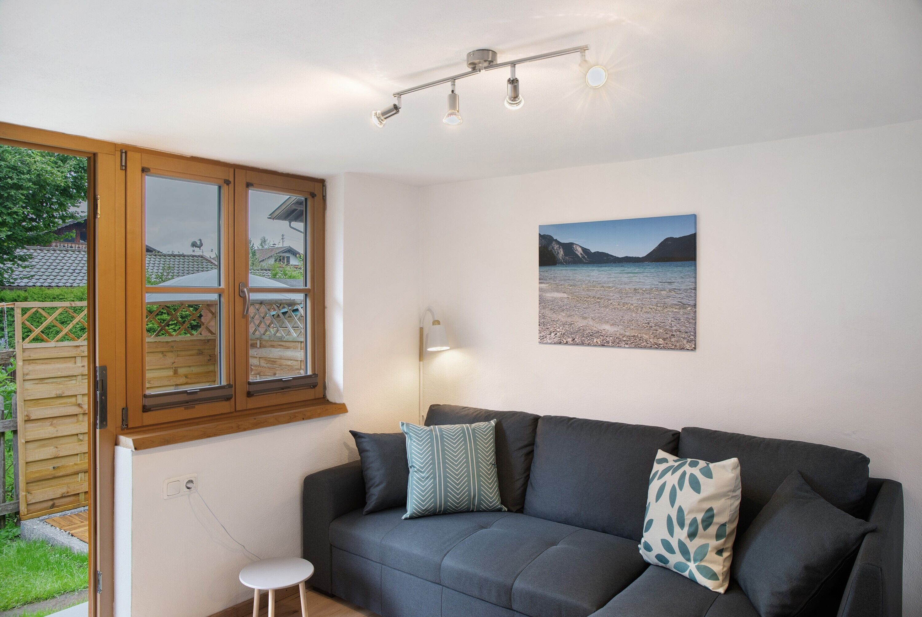 A room with a grey sofa, a small white stool, a window with a view of houses, and a wall-mounted picture.