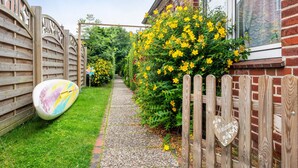 A narrow pathway leading to a house with a wooden fence, a welcome sign, and a surfboard leaning against the fence.