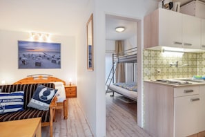 Une pièce compacte comprenant un lit superposé, un petit coin cuisine, une table en bois, un canapé avec des coussins à rayures et un tableau sur le thème de la plage.