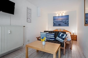 Une pièce comprenant un lit, une table en bois avec un vase de fleurs, un téléviseur fixé au mur, un tableau encadré et un radiateur.