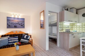 Une pièce comprenant un lit, une petite table avec un vase, un tableau représentant une scène de plage, une kitchenette avec des placards et un miroir.