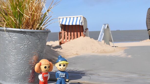 Een strandscène met een potplant, twee figuurtjes, een gestreepte strandparasol en een zandkasteel.
