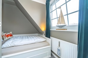 Una camera da letto con soffitto spiovente, un letto con biancheria bianca, una decorazione a forma di barca a vela e una finestra con tende.