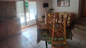 A dining area with wooden chairs and a table, a kitchen with a stone backsplash, and a ceiling with exposed beams.