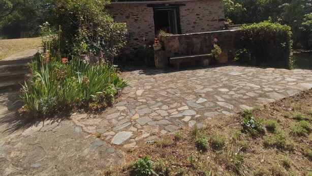 A rustic stone building with a tiled roof, surrounded by greenery and a stone pathway leading to a bench.