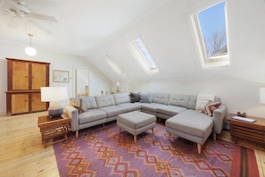 A modern loft bedroom with a large L-shaped sectional sofa, a wooden cabinet, a desk, and a skylight.