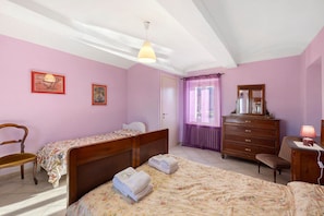 Une chambre avec deux lits simples, une commode en bois, une chaise et un bureau avec une lampe.
