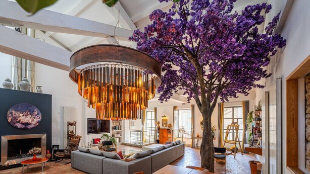 Een ruime woonkamer met een grote bank, een eettafel en een bijzondere boom met paarse bloemen.