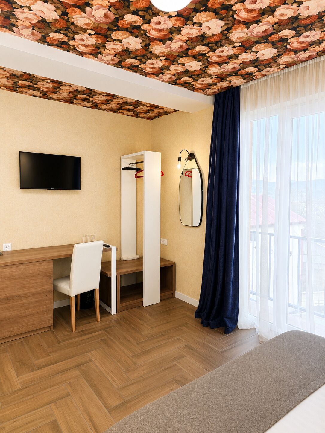 Hotelkamer met een houten visgraatvloer, een flatscreen televisie aan de wand, een bureau met stoel, een grote spiegel en een raam met dunne gordijnen.