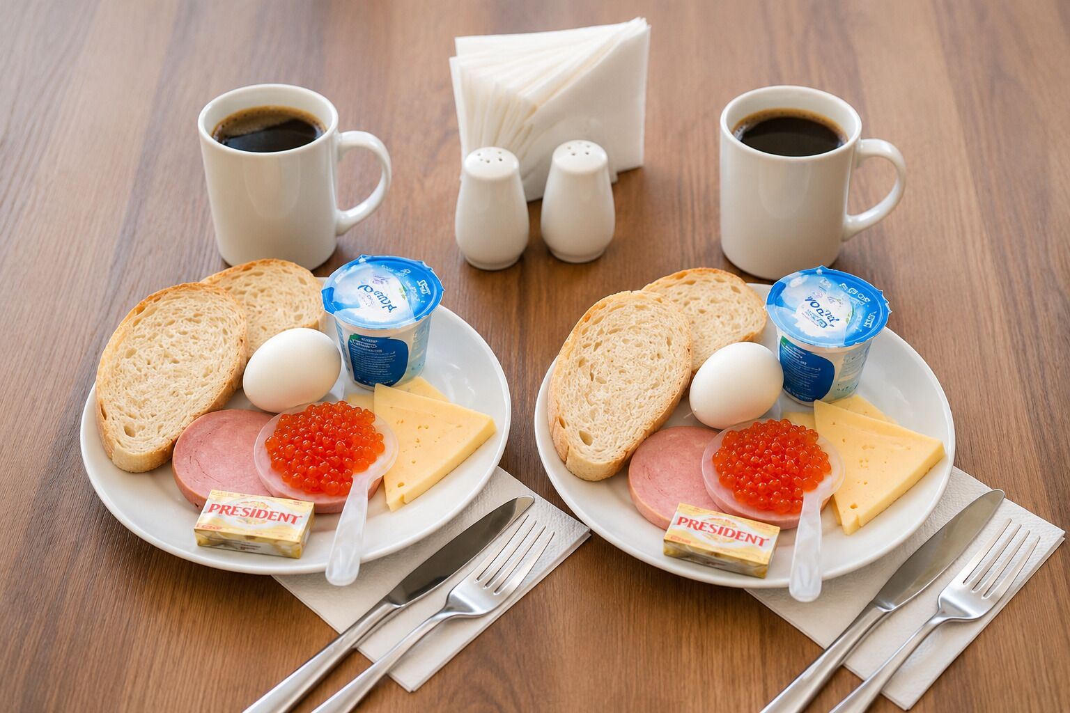 Deux assiettes de nourriture comprenant du pain, du fromage, du jambon, des œufs, du caviar et du yaourt, accompagnées de tasses de café et de couverts sur une table en bois.