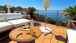 A terrace with a white sofa, a round table, a bottle of wine, and a view of the sea. - Villa Paomia (cargèse)