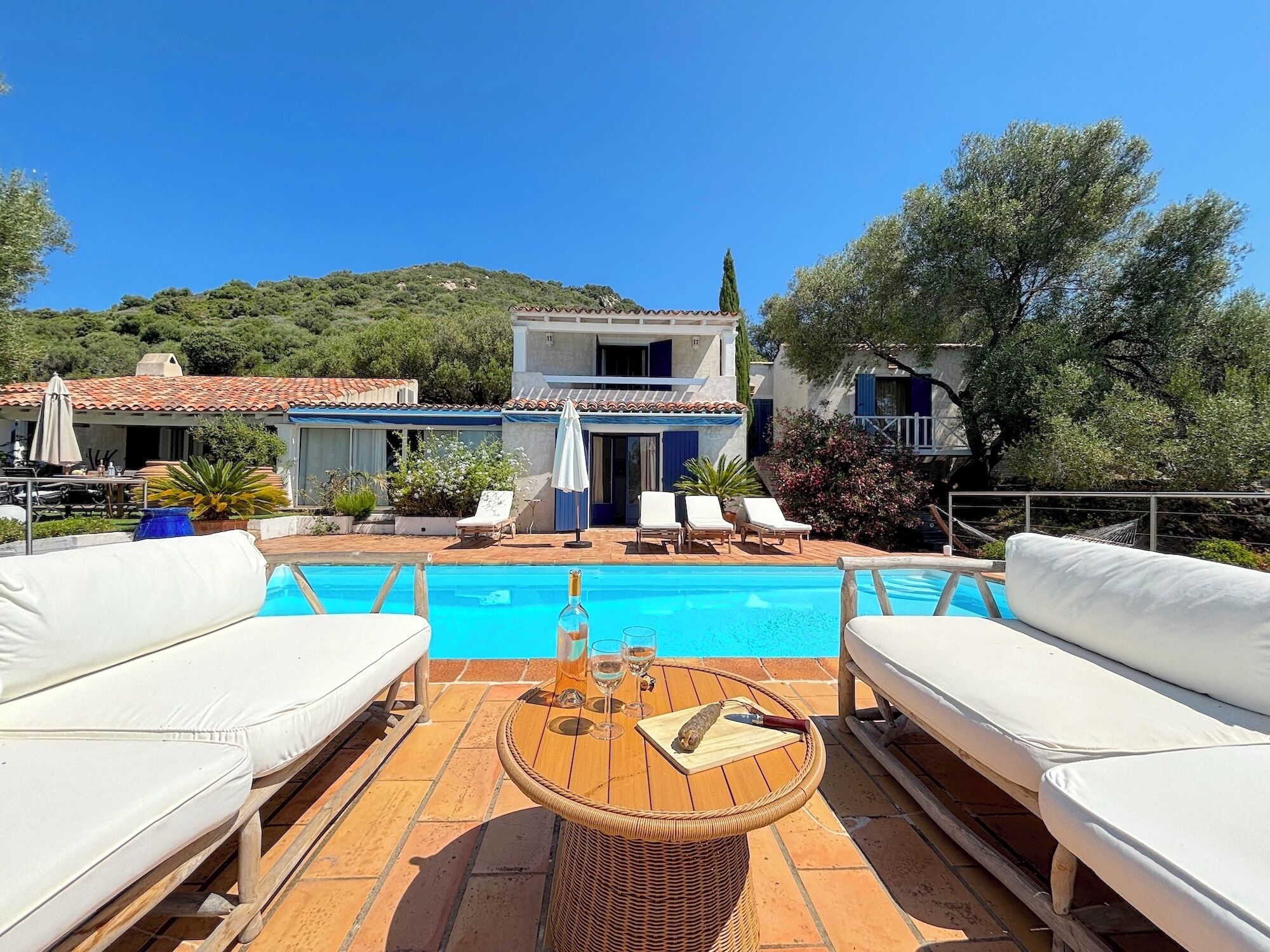 A poolside area with white lounge chairs, a wooden table with a bottle and glasses, and a building with a tiled roof in the background.