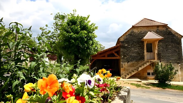 A stone building with a wooden staircase leading to a balcony, surrounded by lush greenery and vibrant flowers.