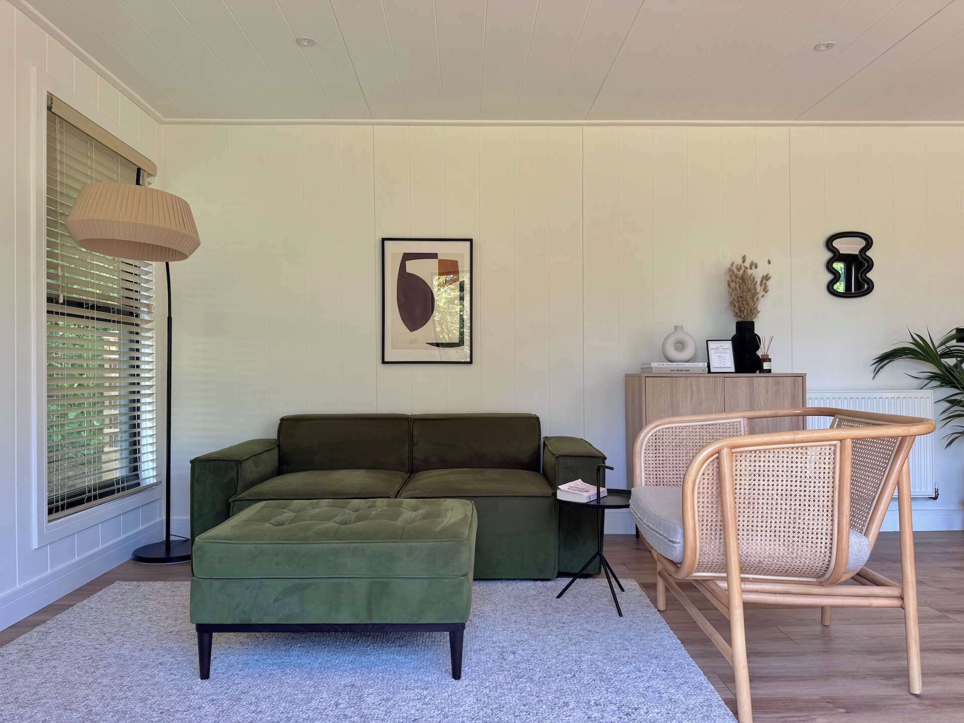 Un soggiorno con un divano verde, un poggiapiedi coordinato, una poltrona in rattan, un'opera d'arte astratta incorniciata e un tavolino con libri e un vaso.