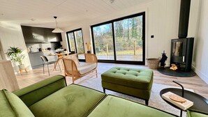 Een moderne woonkamer met groene banken, een rieten stoel, een open haard en een keuken op de achtergrond.