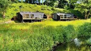 Twee moderne vakantiehuisjes met glazen wanden, gelegen aan een rustige rivier, omgeven door weelderige begroeiing en een veld met klaprozen.